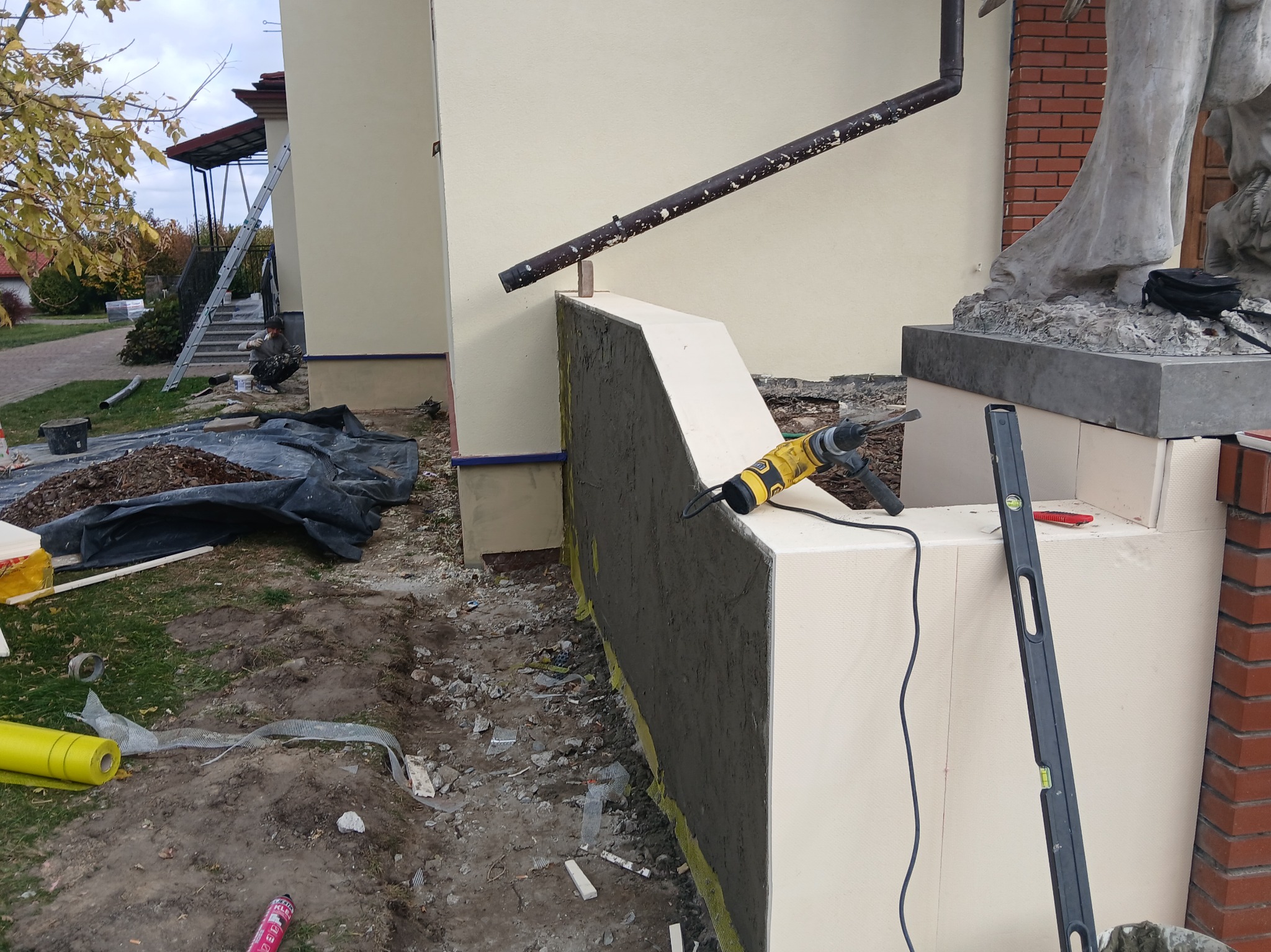 Ocieplanie elewacji: fragment ściany z nałożoną warstwą kleju na siatce, widoczny styropian, poziomica, wiertarka udarowa oraz fragment rynny i rzeźby w tle.