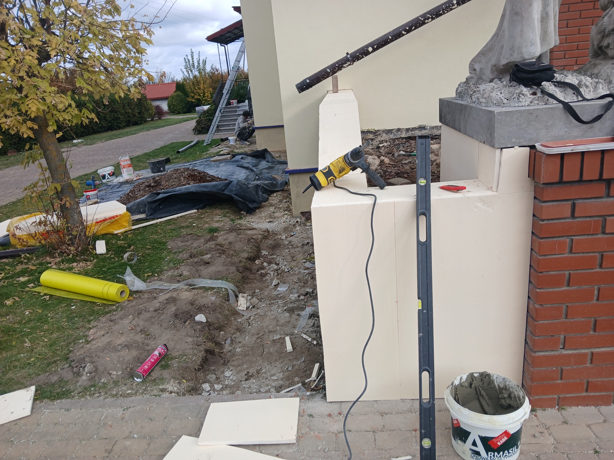 Prace przygotowawcze do wykończenia elewacji budynku: styropianowe płyty, poziomica, wiertarka oraz wiadro z zaprawą murarską na tle domu i ogrodu.