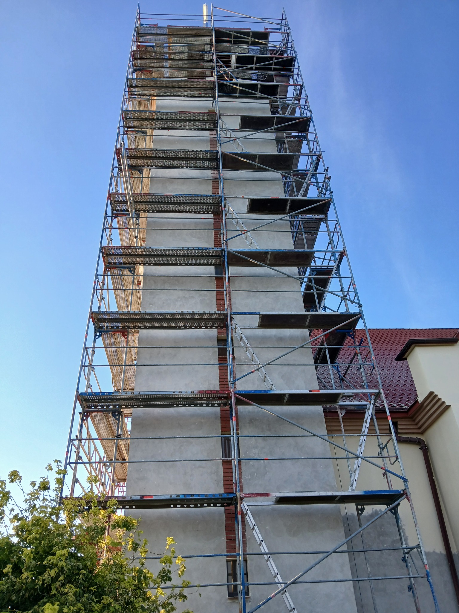 Wysokie rusztowanie otaczające budynek z elewacją w trakcie prac, widoczne fragmenty cegieł i nałożony szary tynk, fragment dachu z czerwonej dachówki w tle, ujęcie z dołu w słoneczny dzień.