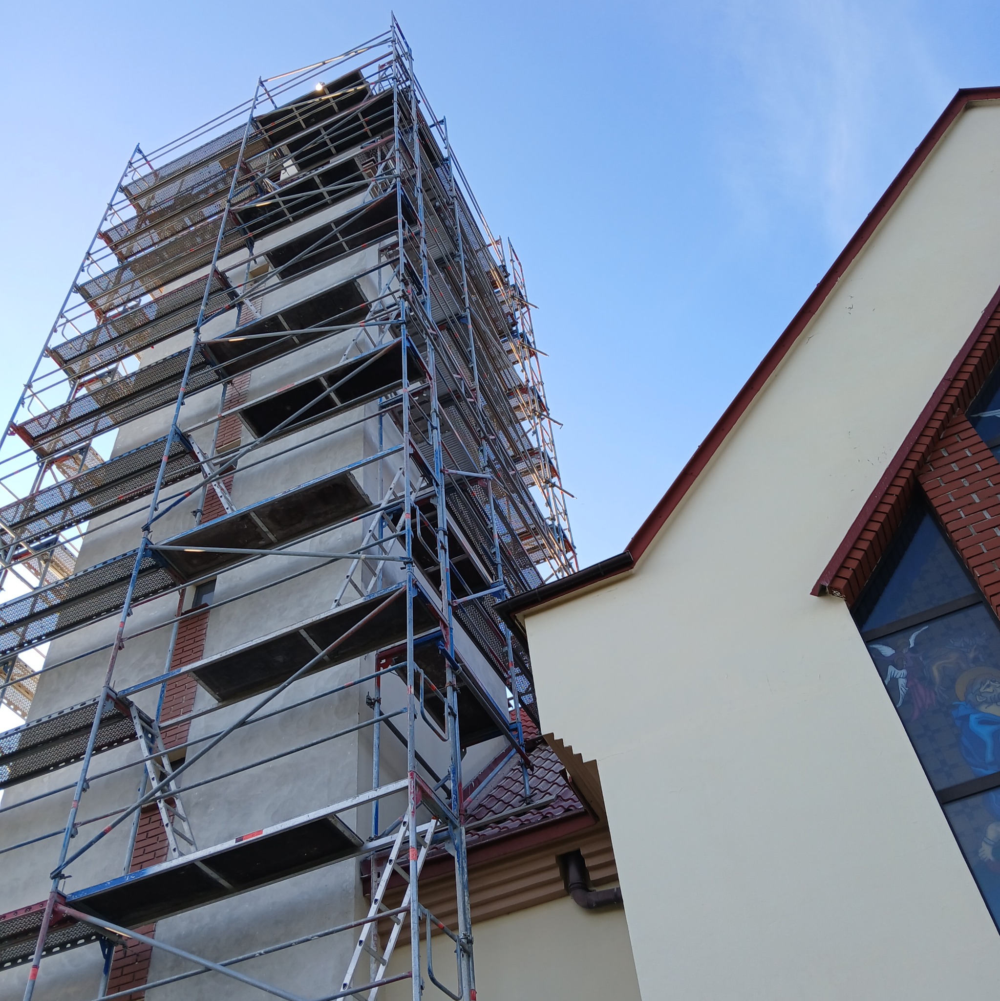Wieża kościoła w trakcie prac budowlanych, otoczona rusztowaniem, widoczny fragment elewacji i okno z witrażem.