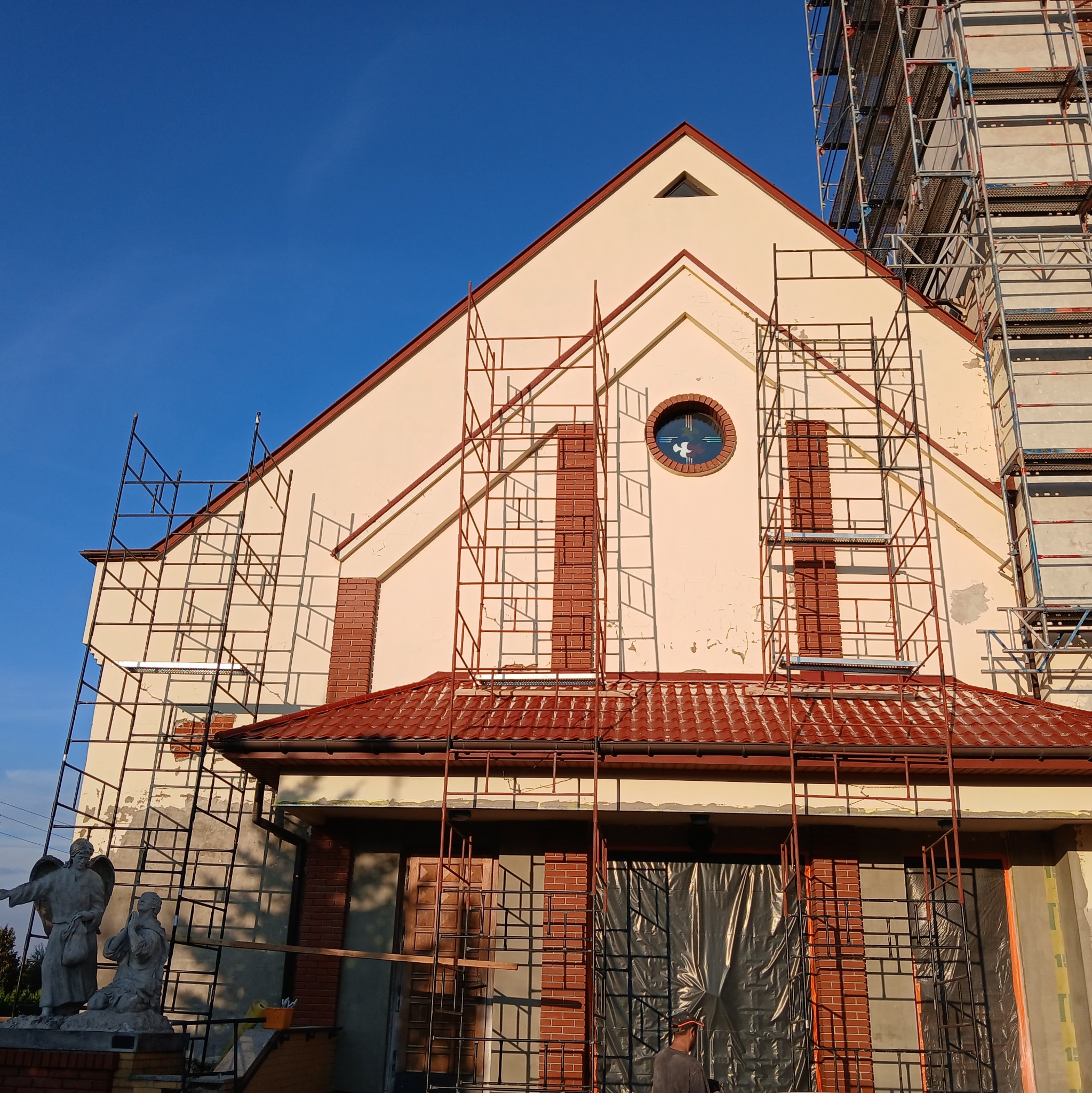 Kościół w trakcie renowacji elewacji, widoczne rusztowania, fragment pomnika oraz fragment elewacji z widocznymi uszkodzeniami i elementami dekoracyjnymi z cegły klinkierowej.