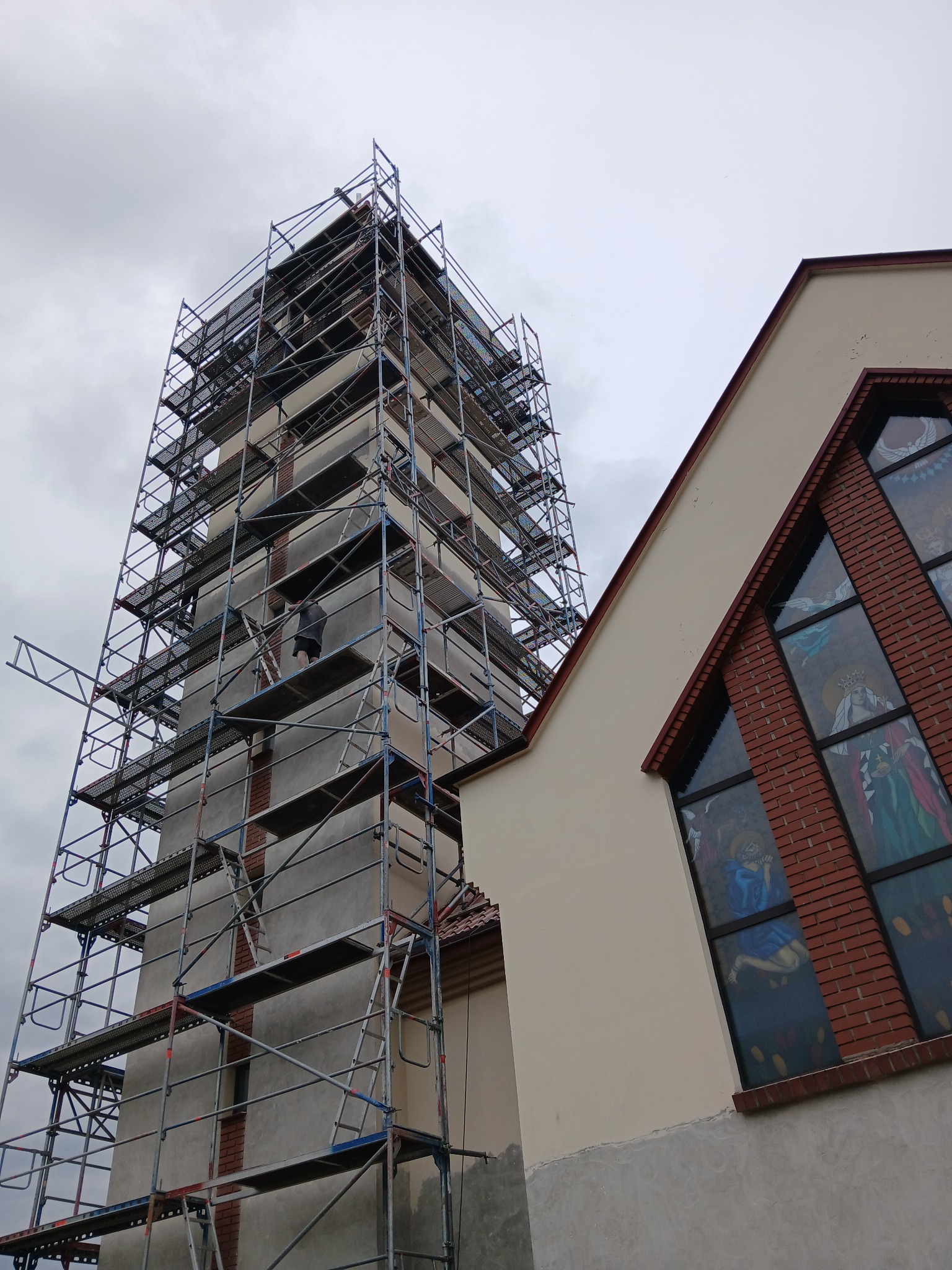 Wysoka wieża kościelna w trakcie prac budowlanych, otoczona metalowym rusztowaniem, widoczne fragmenty elewacji i witraże w oknach. Pracownik na rusztowaniu.