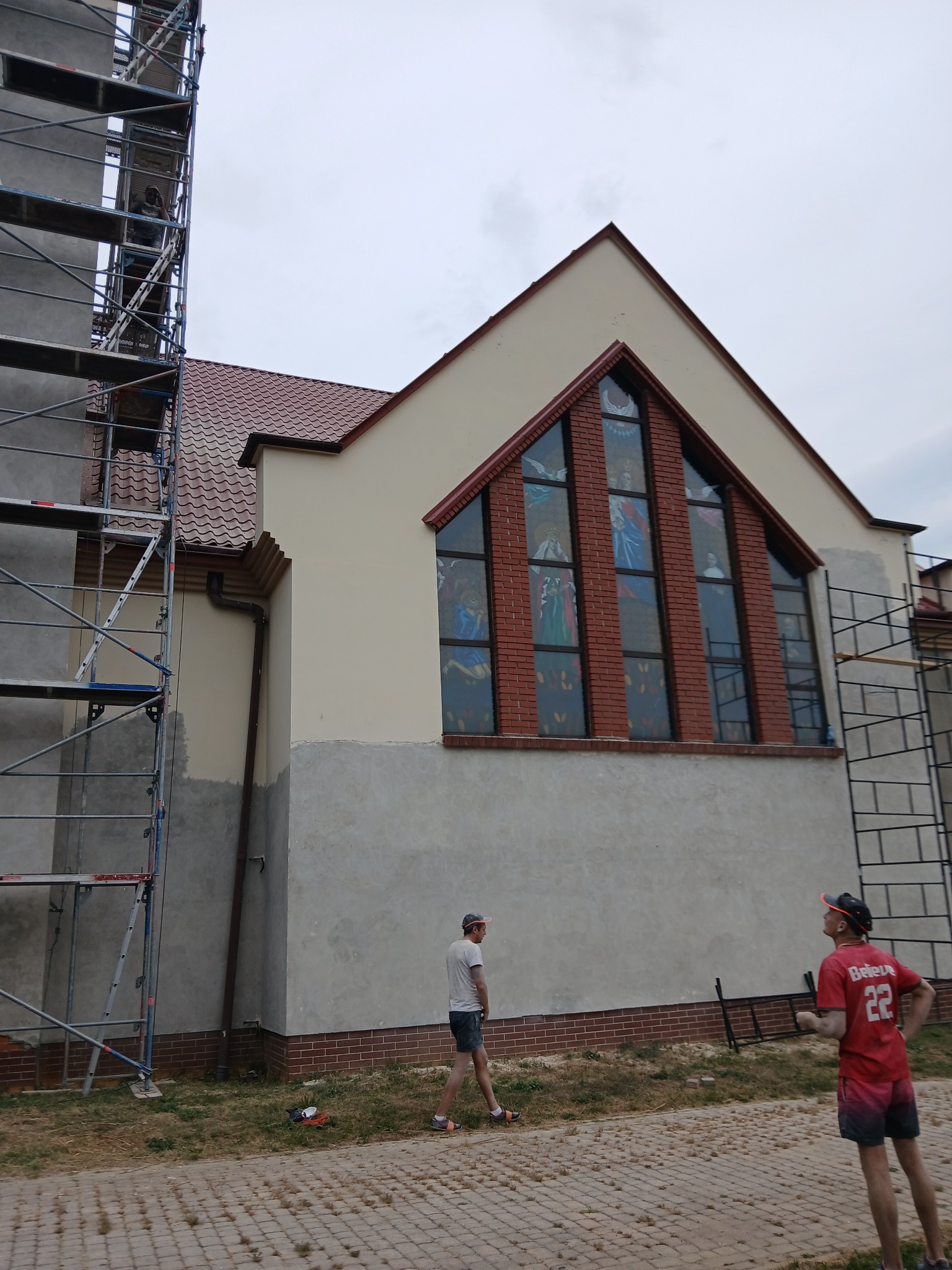 Kościół w trakcie prac elewacyjnych, widoczne rusztowania i fragmenty nieotynkowanej ściany, okna witrażowe z ceglaną obramówką.