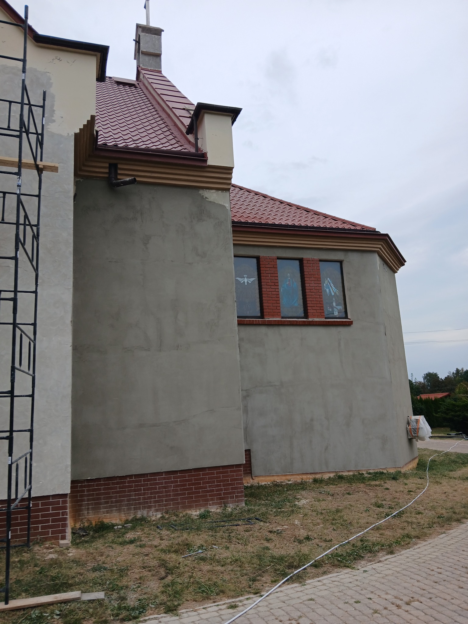 Budynek kościoła w trakcie prac elewacyjnych, z widocznym rusztowaniem, świeżo otynkowaną ścianą i fragmentem dachu z czerwoną dachówką.