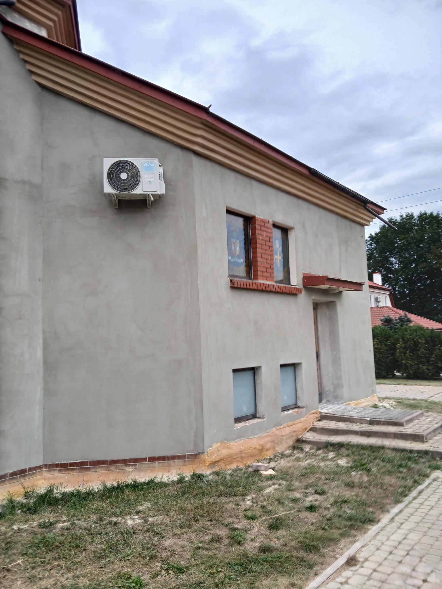 Ściana budynku po nałożeniu szarego tynku w Piasecznie, widoczny klimatyzator na elewacji i ceglana podmurówka.