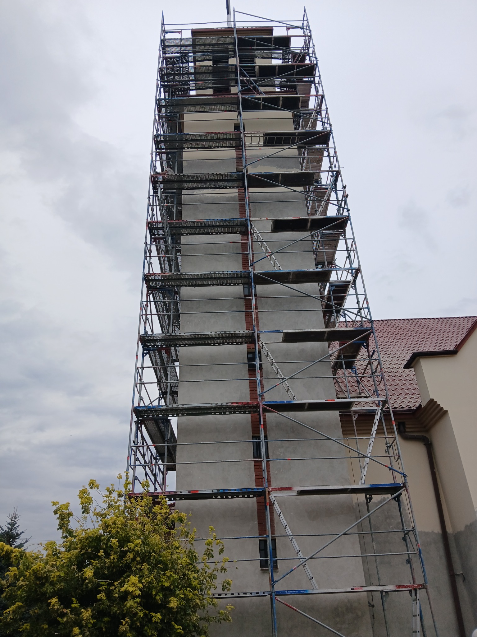Wysoka wieża budynku w trakcie prac, otoczona rusztowaniem, widok z dołu na pochmurne niebo.