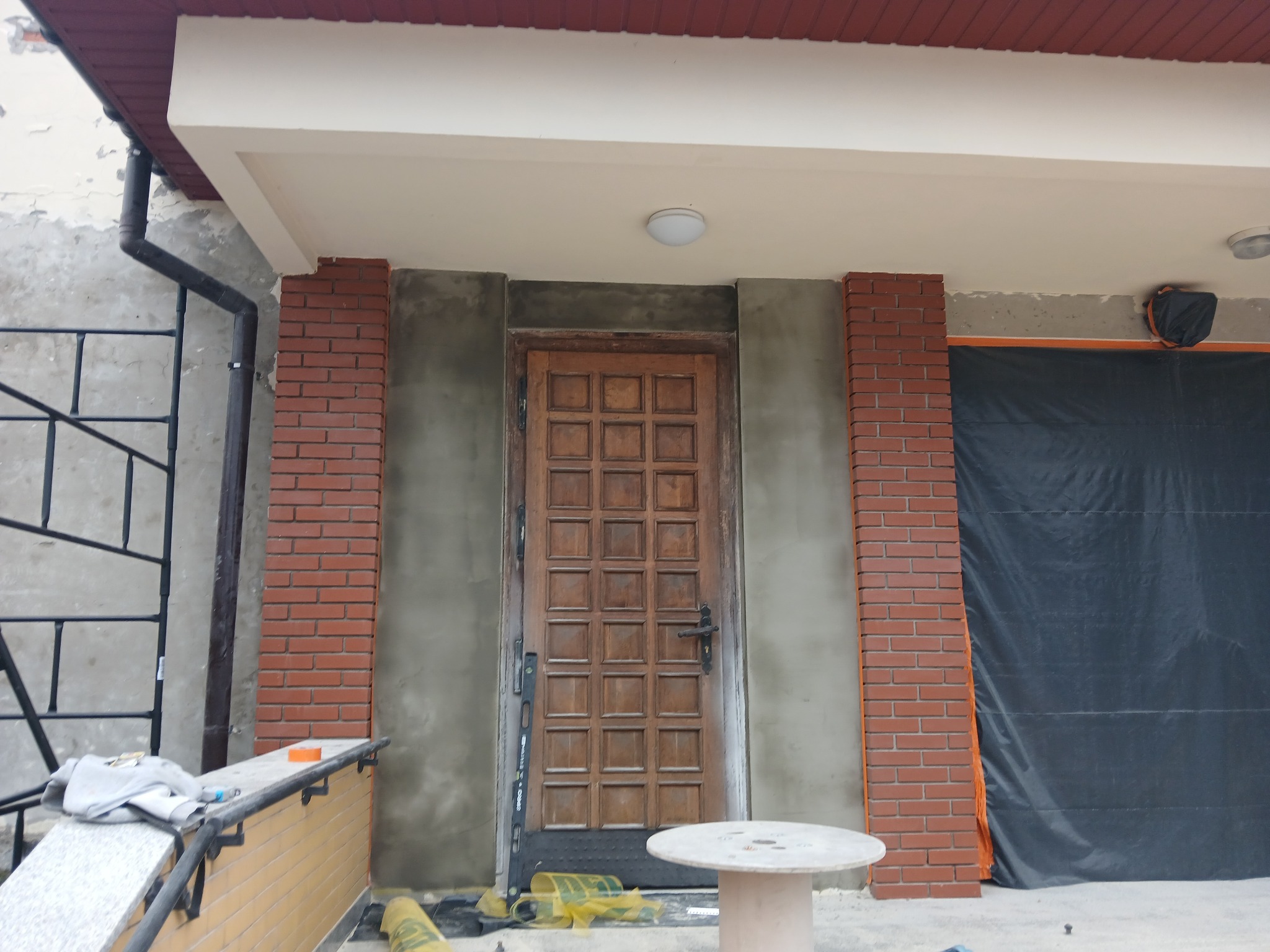 Wejście do domu w trakcie prac: otynkowana ściana, ceglane pilastry, drewniane drzwi wejściowe z kratownicą, osłonięty fragment elewacji folią ochronną, widoczne narzędzia i materiały budowlane.