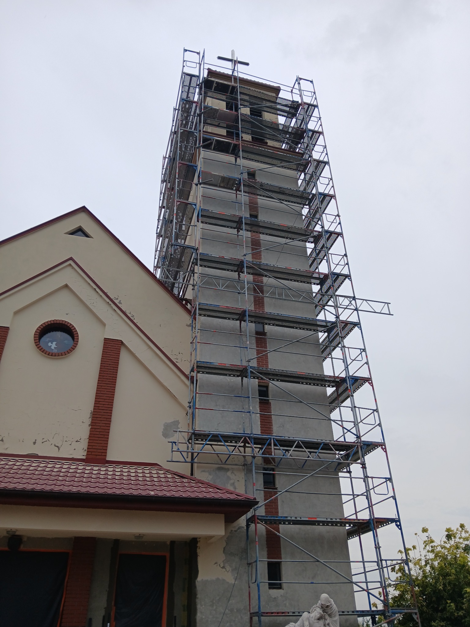 Wieża kościoła w trakcie renowacji, otoczona rusztowaniem, z widocznymi śladami napraw elewacji i krzyżem na szczycie.