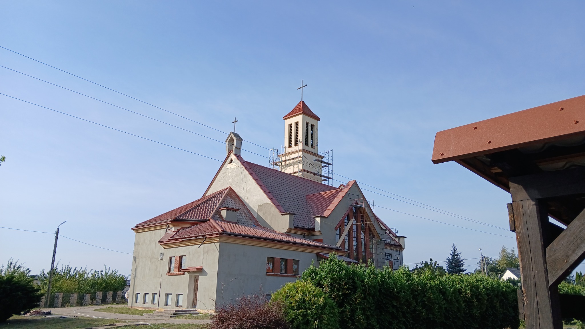 Kościół w trakcie prac remontowych; widoczne rusztowania wokół wieży i na elewacji, nowa czerwona dachówka, szara elewacja w trakcie wykończenia.