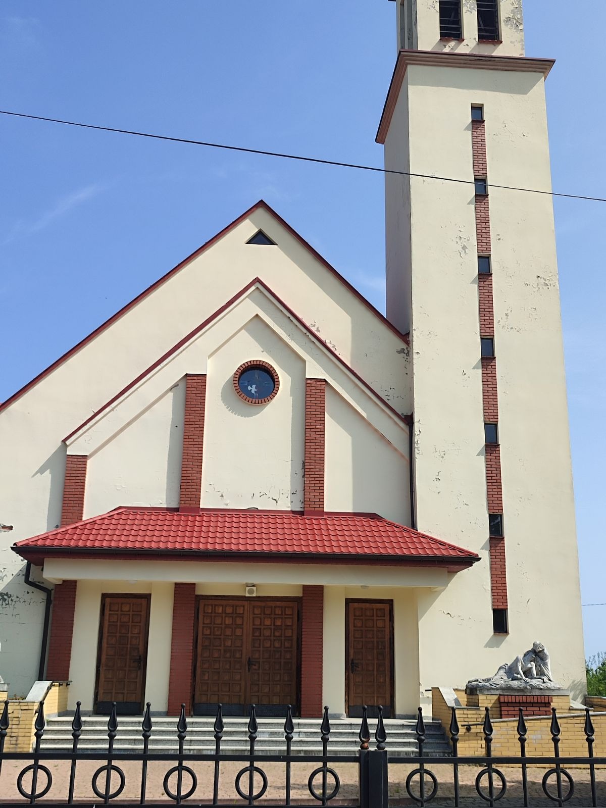 Kościół z kremową elewacją, czerwoną dachówką i wysoką wieżą, widoczne ślady zabrudzeń i łuszczącej się farby na ścianach, żelazne ogrodzenie na pierwszym planie.