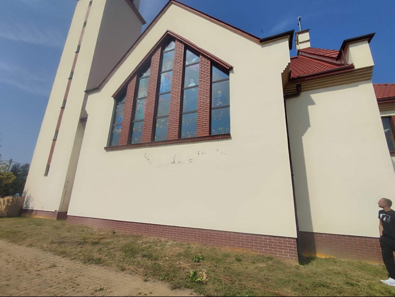 Kościół z beżową elewacją, czerwonym dachem i witrażowymi oknami, na ścianie widoczne ślady po usuniętych napisach, na pierwszym planie trawa i chodnik, w tle błękitne niebo.