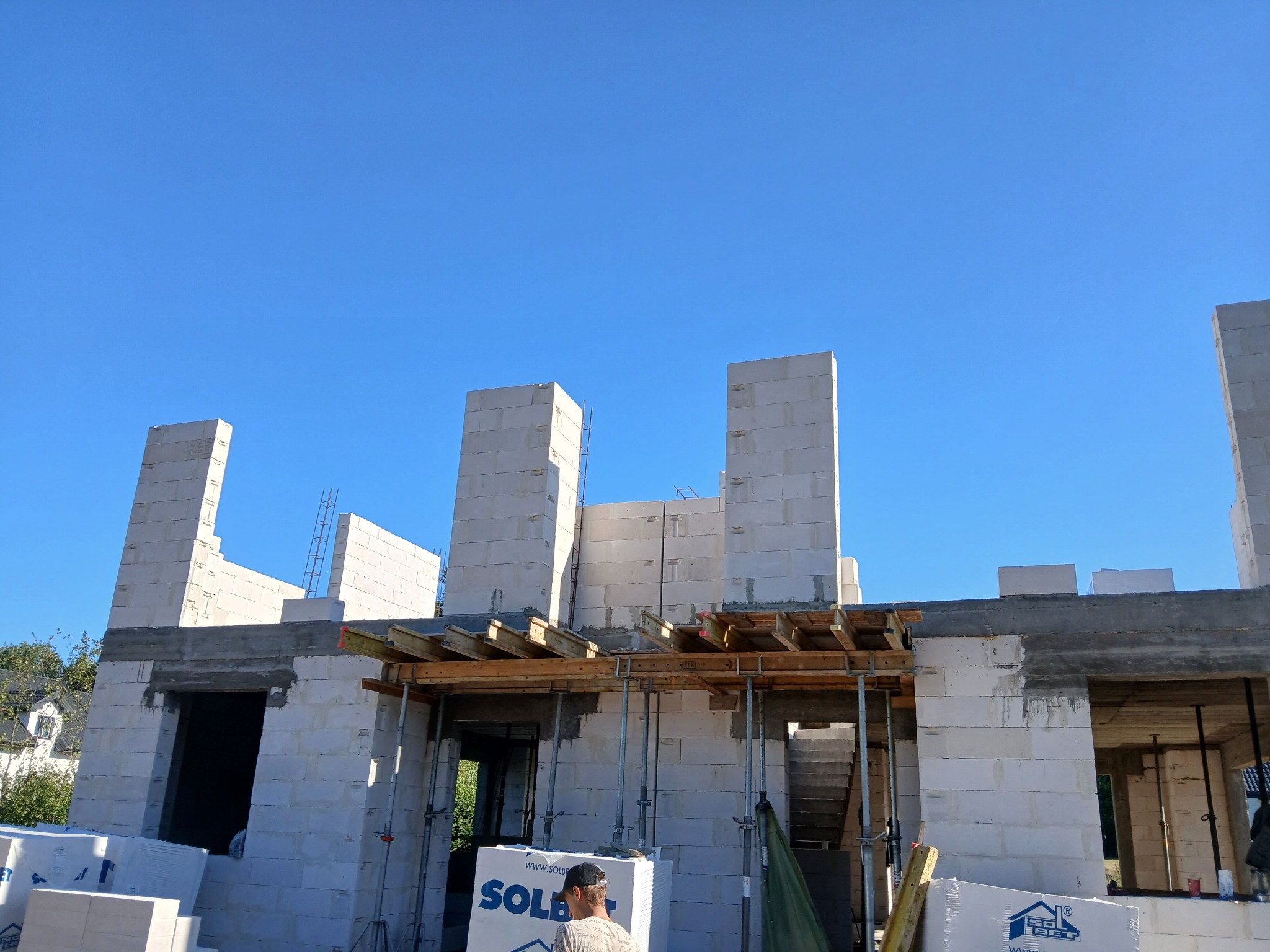 Budowa domu w trakcie wznoszenia ścian z białych bloczków, widoczne drewniane szalunki i zbrojenia, fragment osoby w dolnej części kadru, błękitne niebo w tle.