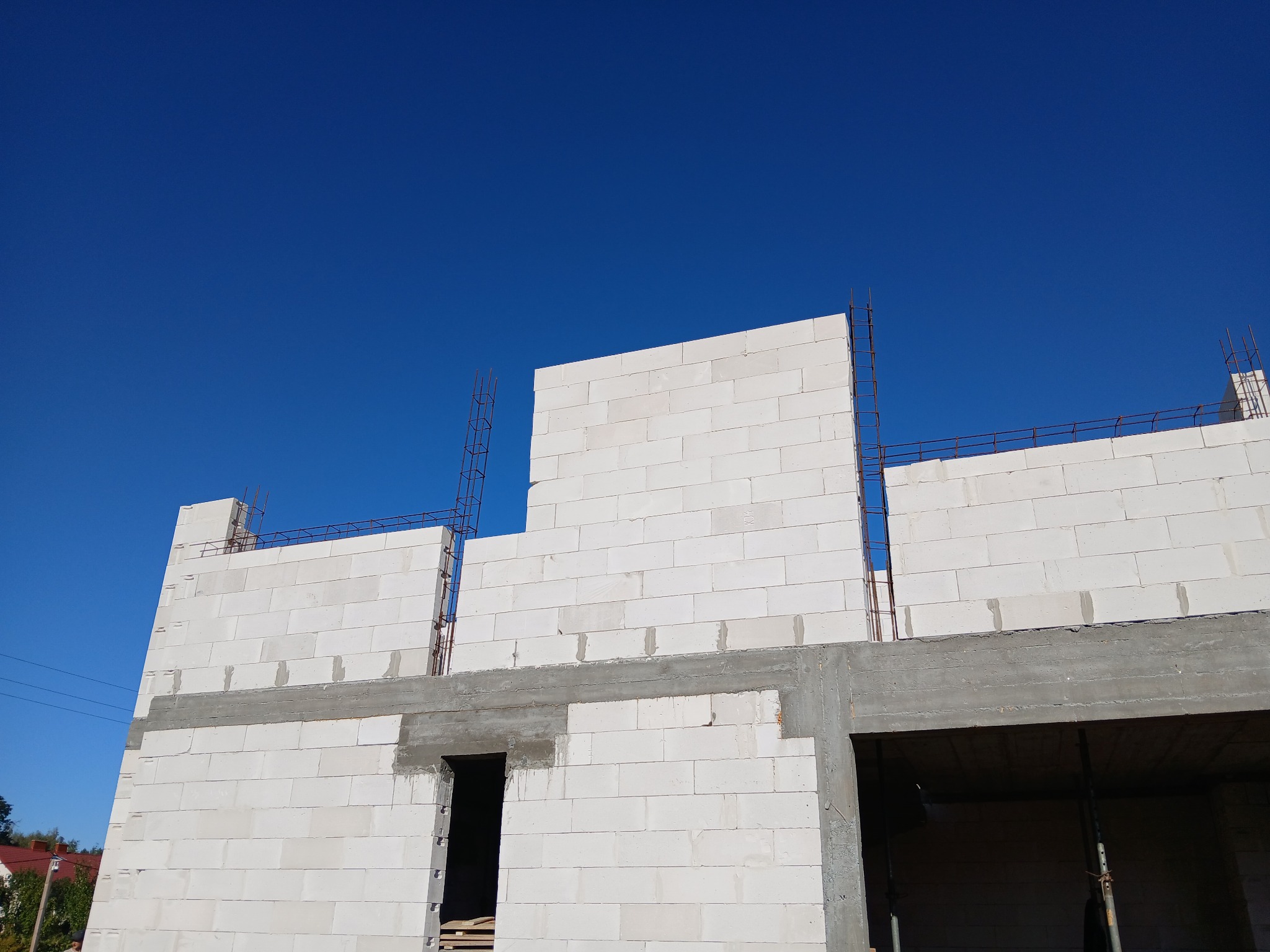 Wznoszenie ścian domu z białych bloczków betonowych, widoczne zbrojenie i otwory okienne na tle błękitnego nieba.