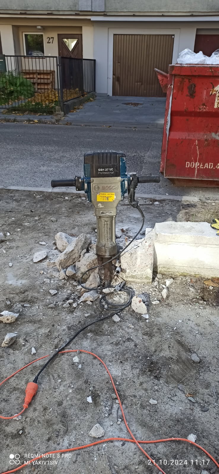 Profesjonalny młot udarowy Bosch GSH 27 VC wbity w gruz na tle budynku z numerem 27 i czerwonym kontenerem na śmieci.