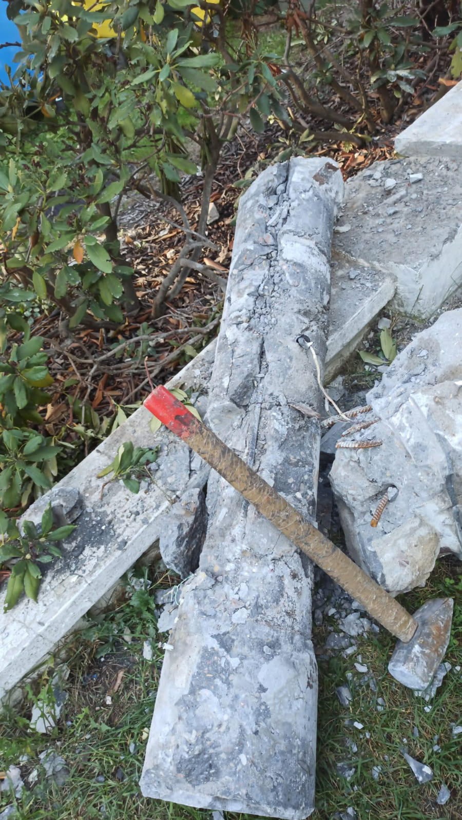 Fragmenty gruzu betonowego, w tym kolumna z widocznym zbrojeniem, leżące na trawie obok krzewu. Obok gruzu stoi młotek z czerwoną końcówką trzonka.