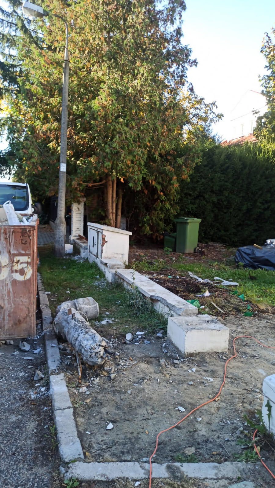 Teren po rozbiórce z gruzem, fragmentami fundamentów, kontenerem na odpady i zielenią w tle.