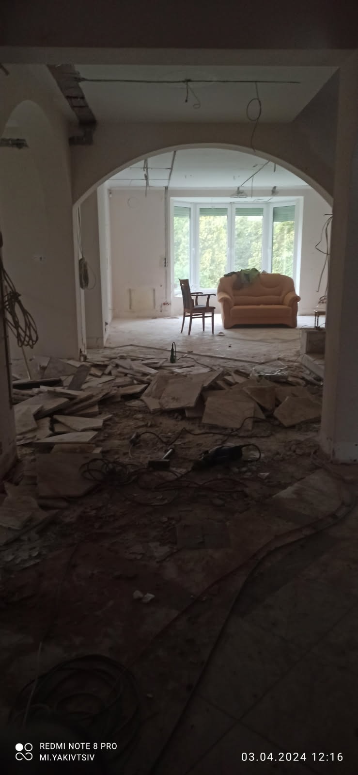 Wnętrze domu w trakcie rozbiórki: gruz na podłodze, widoczne przewody elektryczne, częściowo usunięta ściana łukowa, w tle okno z widokiem na drzewa, fotel i kanapa.