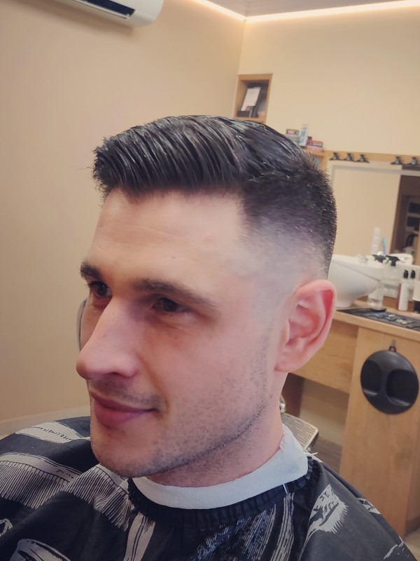 Mężczyzna z krótką, stylowo ułożoną fryzurą z wyraźnym cieniowaniem, widok z profilu w salonie z jasnym oświetleniem.