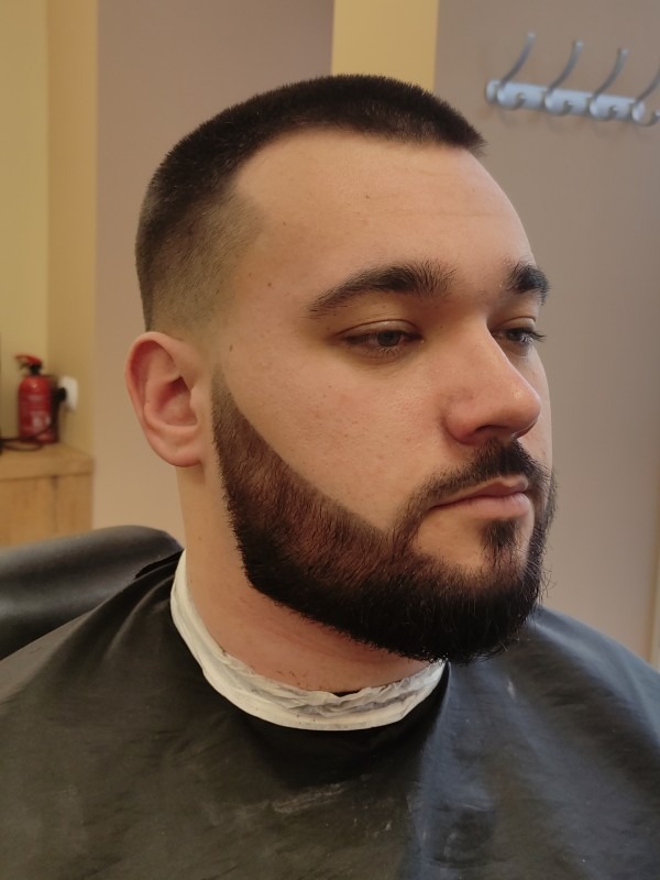 Mężczyzna z krótką fryzurą i precyzyjnie wystylizowaną brodą w salonie w Warszawie, widziany z profilu.