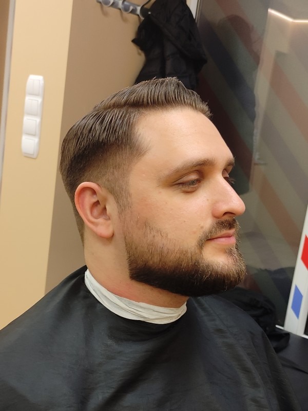 Mężczyzna z modną fryzurą i starannie przystrzyżoną brodą, widziany z profilu w salonie fryzjerskim, ubrany w pelerynę ochronną.