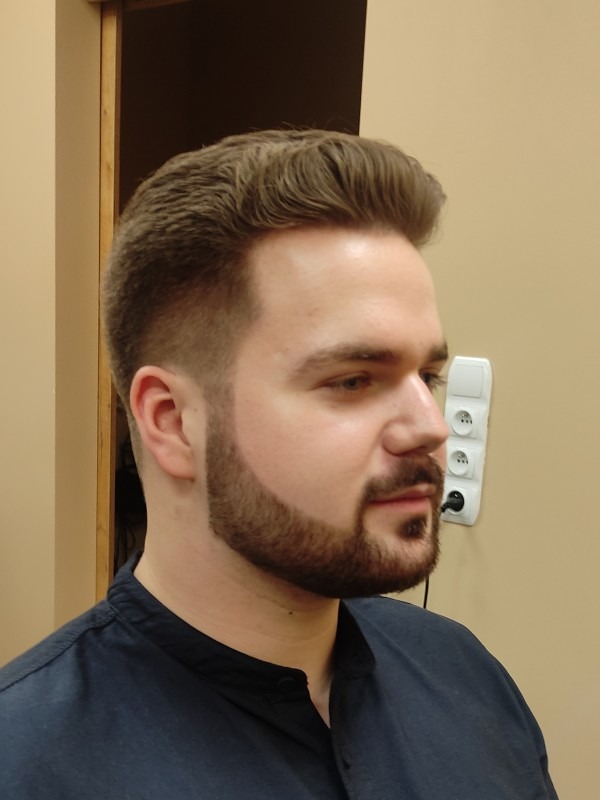 Mężczyzna z modną fryzurą i starannie przystrzyżoną brodą, widok z profilu na tle beżowej ściany z gniazdkiem elektrycznym.