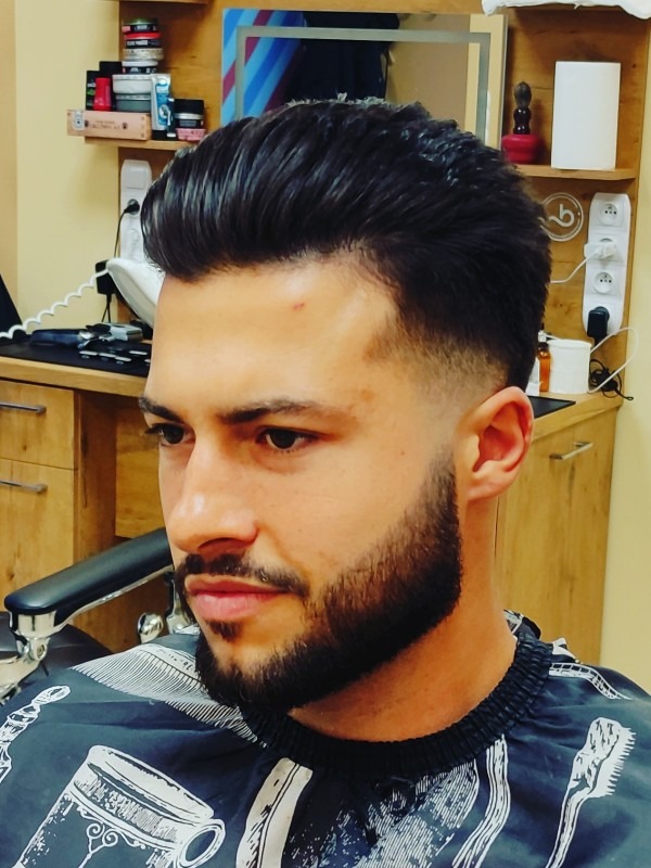 Mężczyzna z modną fryzurą typu pompadour i starannie przystrzyżoną brodą, w pelerynie ochronnej z wzorem, po zabiegu stylizacji włosów.