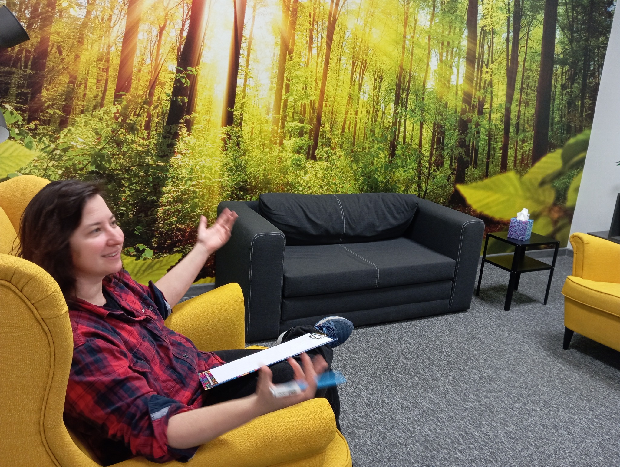 Kobieta w czerwonej koszuli w kratę siedzi w żółtym fotelu na tle fototapety z motywem leśnym, gestykulując rękami podczas rozmowy w gabinecie z szarą sofą i stolikiem z chusteczkami.