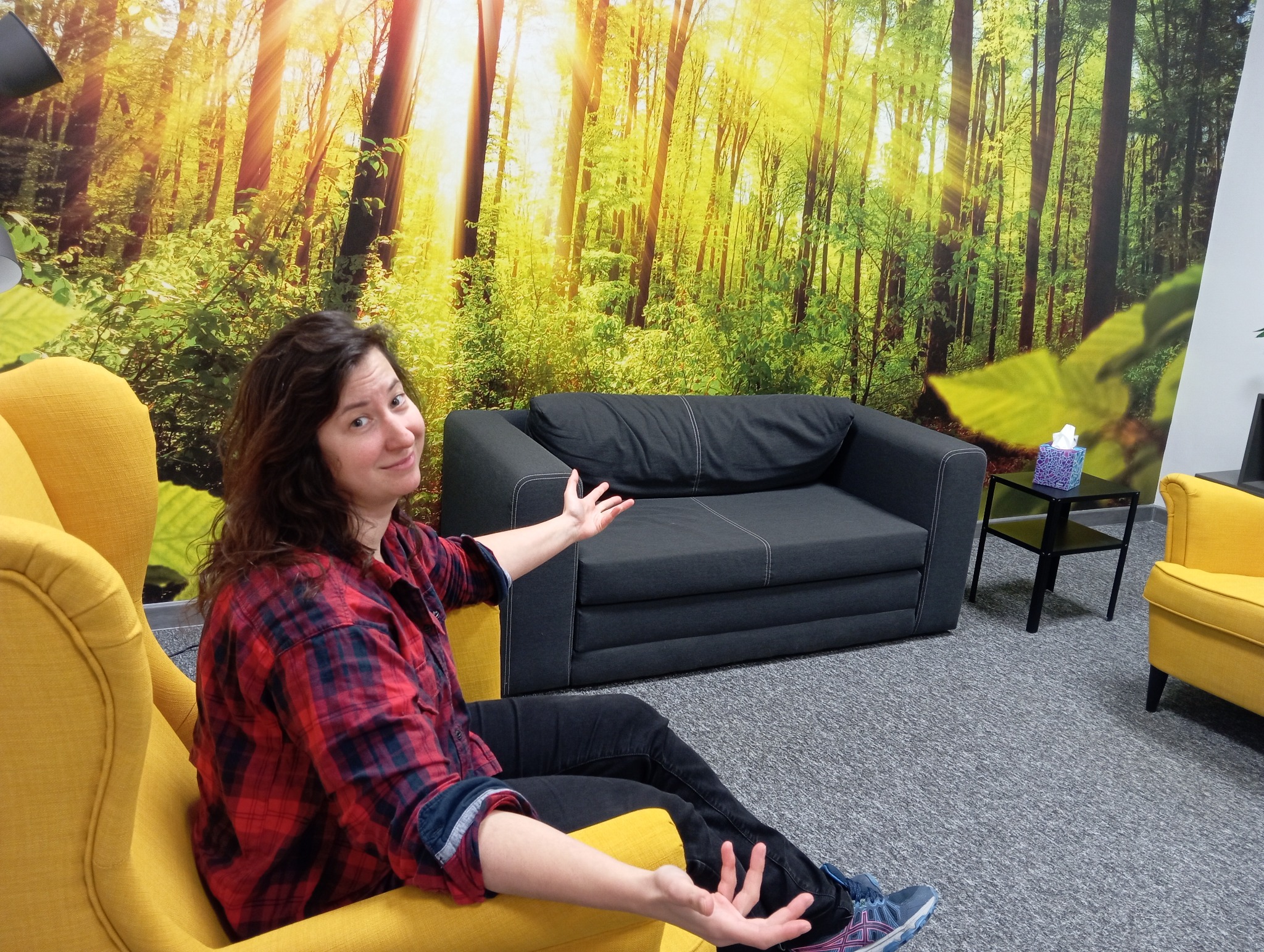 Kobieta w czerwonej koszuli w kratę siedzi w żółtym fotelu na tle fototapety z lasem, gestykulując rękami w kierunku szarej sofy.