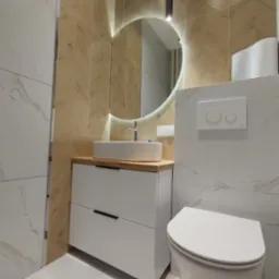 Nowoczesna łazienka z umywalką nablatową na białej szafce z drewnianym blatem, podświetlane okrągłe lustro, wisząca toaleta i płytki imitujące marmur oraz drewno w jodełkę.