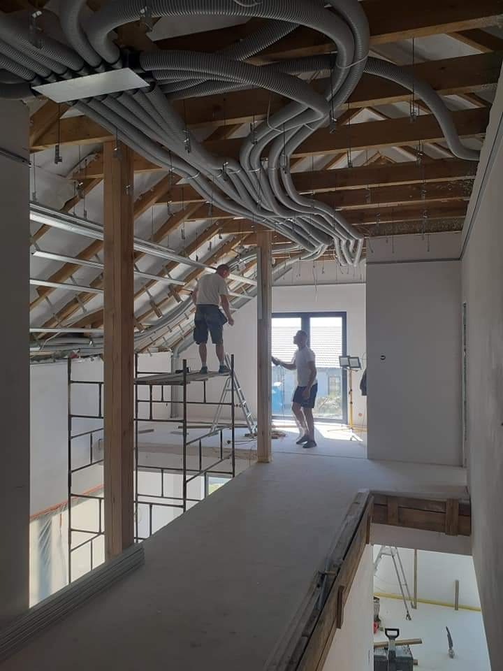 Montaż rur wentylacyjnych pod sufitem na poddaszu, widoczni dwaj pracownicy na rusztowaniu i drabinie, surowe wnętrze w trakcie wykańczania.