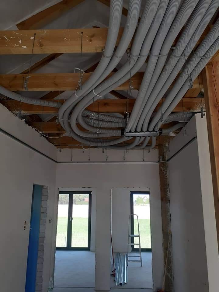 Wnętrze w stanie surowym z widocznymi szarymi, karbowanymi rurami instalacji elektrycznej podwieszonymi do drewnianej konstrukcji stropu. Na podłodze leżą profile metalowe, w tle widoczne okna...