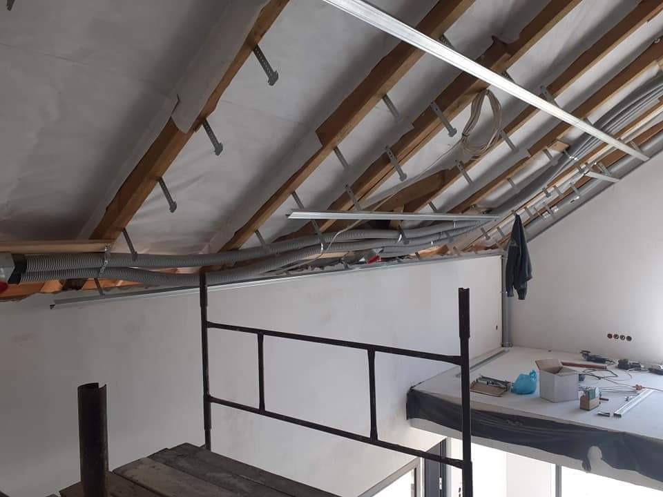 Poddasze w trakcie adaptacji: widoczne drewniane krokwie, izolacja, rury instalacyjne w peszlach, metalowe profile sufitowe, rusztowanie i przygotowane stanowisko pracy z narzędziami.