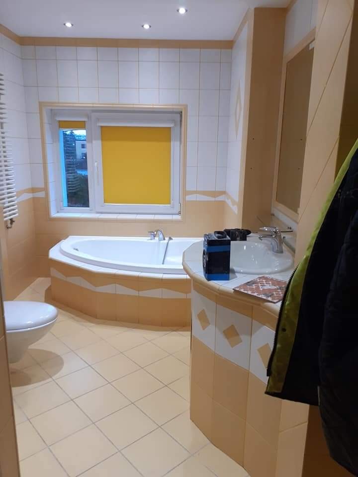 Ukończona łazienka z wanną narożną obudowaną płytkami w odcieniach beżu i bieli, widoczny żółty roleta w oknie i narzędzia na blacie umywalki.