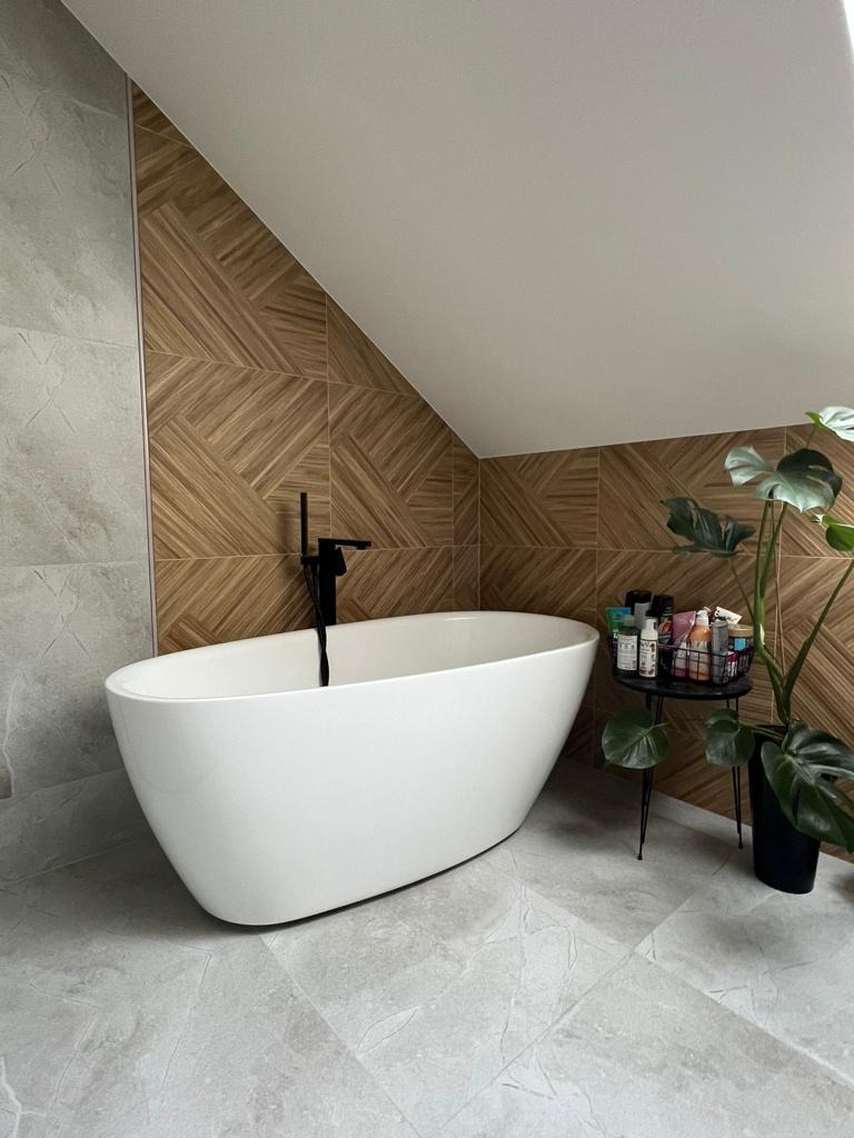 Wolnostojąca biała wanna z czarną armaturą na tle ściany wyłożonej drewnopodobnymi płytkami w geometryczny wzór, uzupełniona szarą płytką podłogową i rośliną doniczkową.