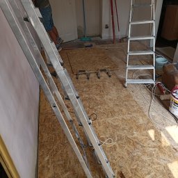 PiotrBodetko - Ściana po wstępnym wyrównaniu, widoczna aluminiowa drabina, podłoga wyłożona płytami OSB, wiadro z farbą Śnieżka, narzędzia malarskie i pędzel.