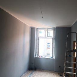 PiotrBodetko - Pokój po świeżym pomalowaniu ścian na szaro, z białym sufitem i listwami, drabina oparta o ścianę, okno otwarte, widoczne narzędzia malarskie.