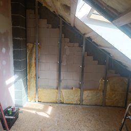 PiotrBodetko - Poddasze w trakcie ocieplania: widoczne wełna mineralna między metalowymi profilami, bloczki betonowe, okno dachowe i narzędzia na podłodze z płyt OSB.