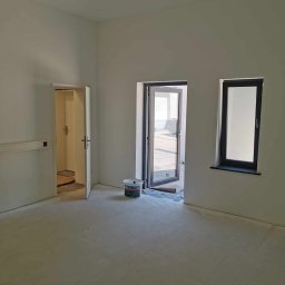 PiotrBodetko - Wnętrze pokoju po remoncie, świeżo pomalowane ściany na biało, widoczne drzwi i okno z czarnymi ramami, na podłodze ślady farby, wiaderko farby marki Brillux.