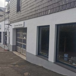 PiotrBodetko - Odnowiona elewacja budynku usługowego z szarą dachówką, widoczne rusztowanie i logo 'Economy Service', dolna część ściany pomalowana na szaro.