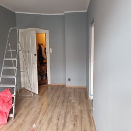 PiotrBodetko - Świeżo pomalowane na szaro wnętrze pokoju z widoczną drabiną, workiem na śmieci i otwartymi drzwiami do innego pomieszczenia.