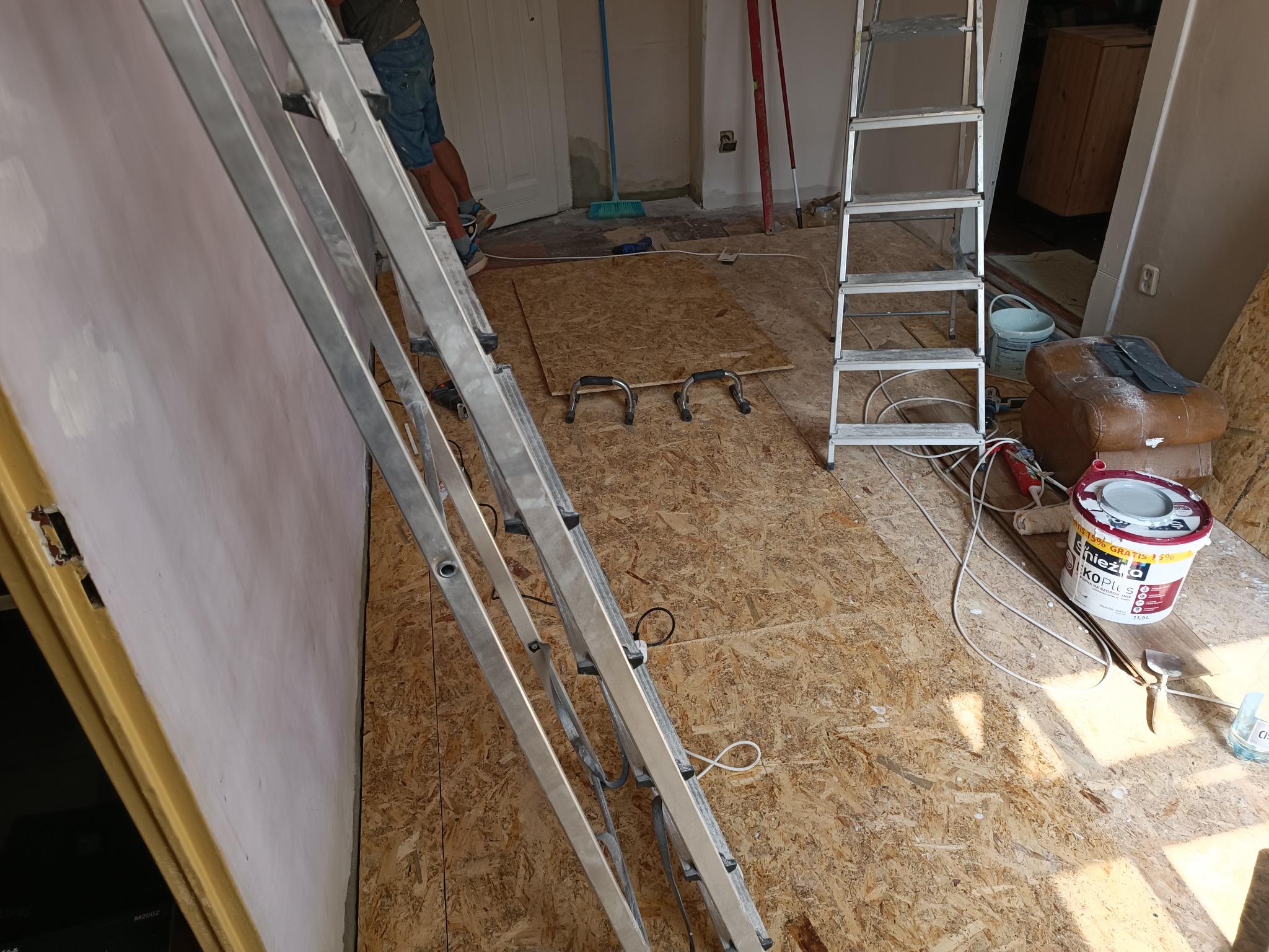 Ściana po wstępnym wyrównaniu, widoczna aluminiowa drabina, podłoga wyłożona płytami OSB, wiadro z farbą Śnieżka, narzędzia malarskie i pędzel.