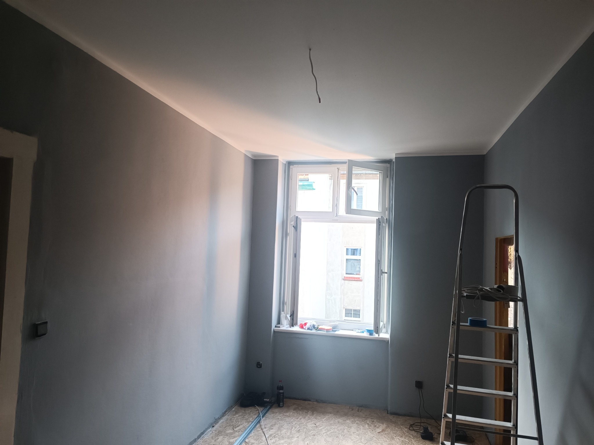 Pokój po świeżym pomalowaniu ścian na szaro, z białym sufitem i listwami, drabina oparta o ścianę, okno otwarte, widoczne narzędzia malarskie.