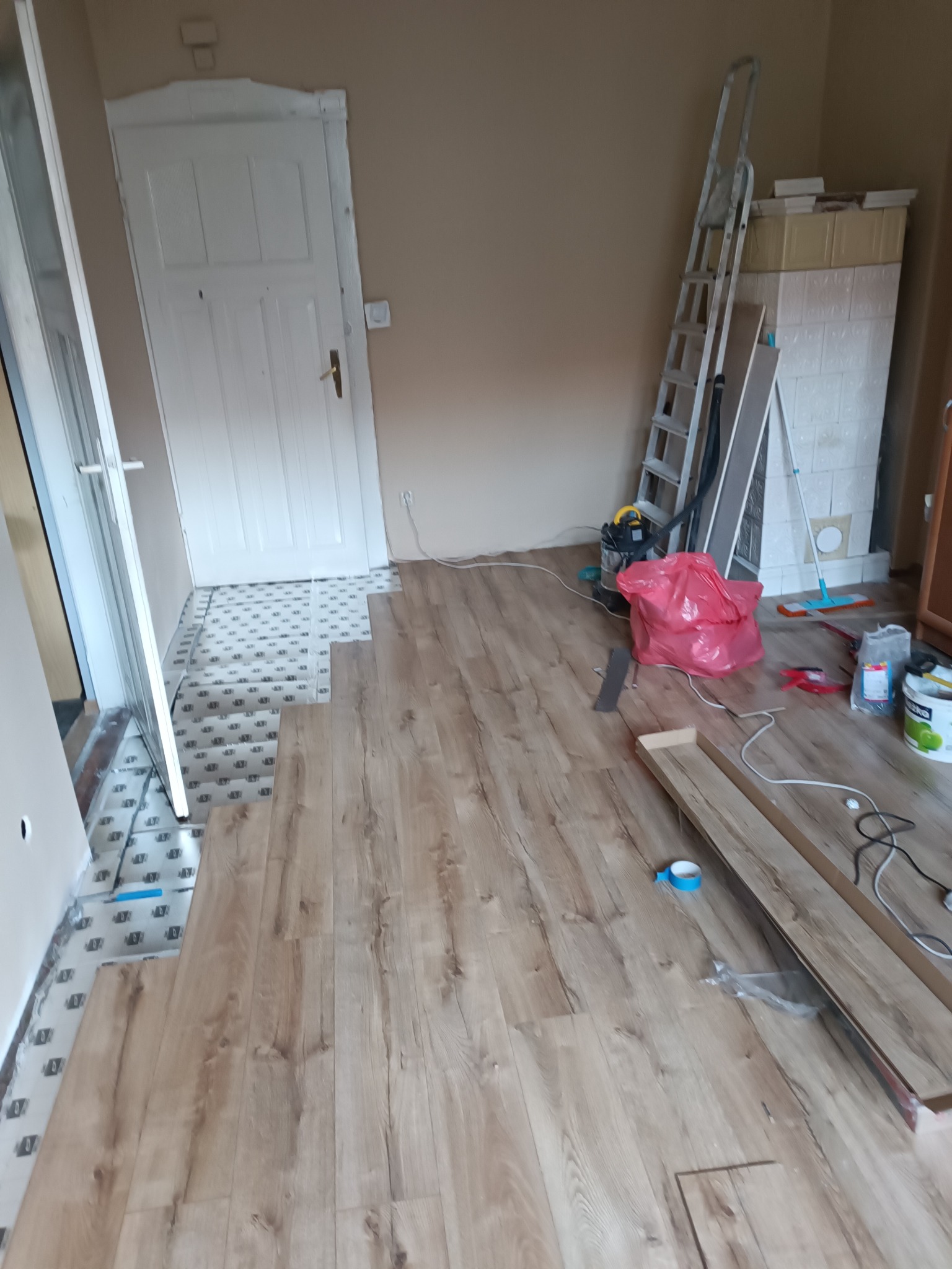 Układanie paneli podłogowych w jasnym odcieniu drewna, widoczna podkładka izolacyjna, białe drzwi w tle, drabina oparta o piec kaflowy, czerwony worek na gruz i narzędzia pracy.