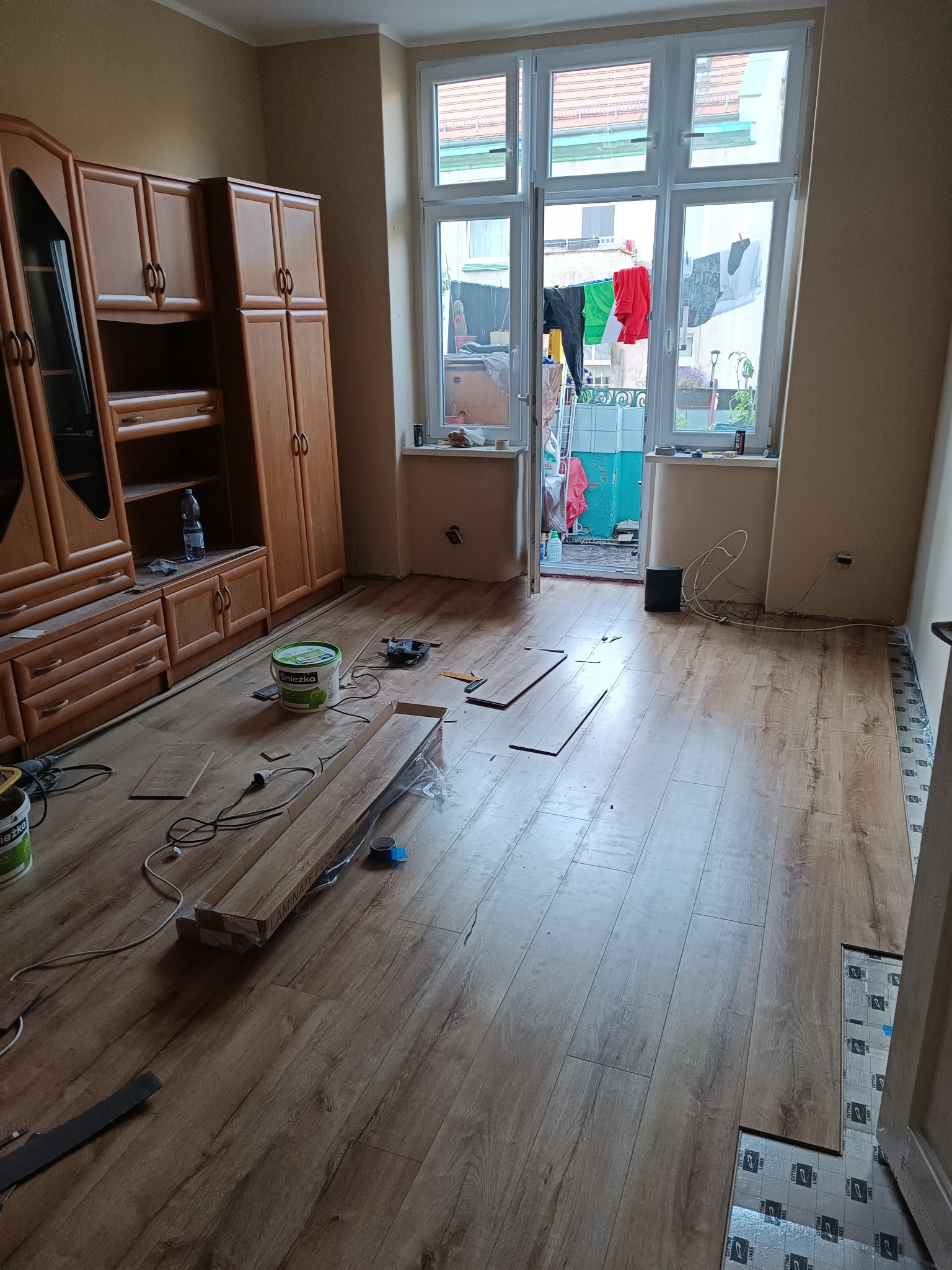 Układanie paneli podłogowych w pokoju z widocznymi narzędziami, paczką paneli, puszkami z farbą Śnieżka i częściowo ułożoną podłogą na podkładzie izolacyjnym.