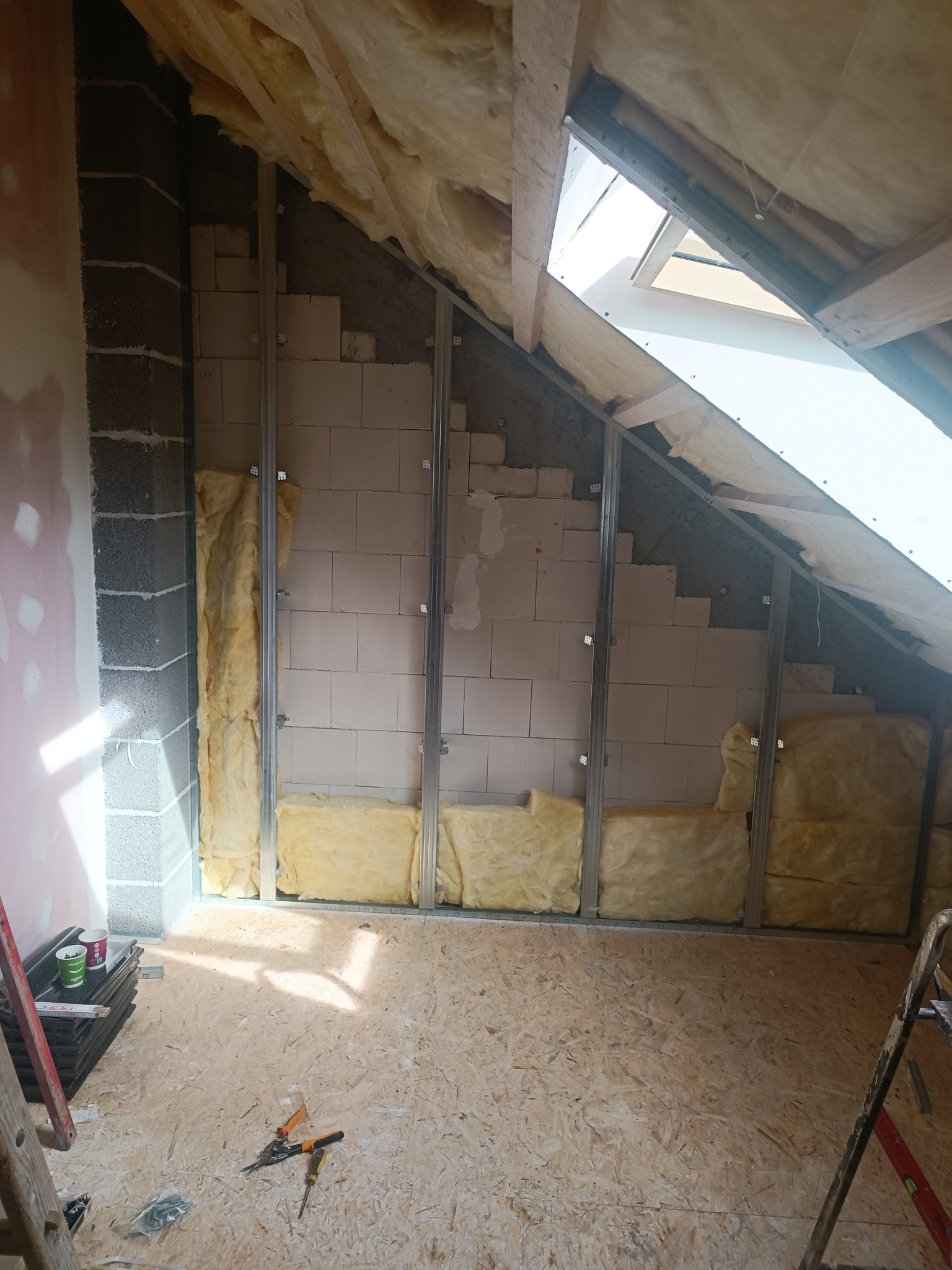 Poddasze w trakcie ocieplania: widoczne wełna mineralna między metalowymi profilami, bloczki betonowe, okno dachowe i narzędzia na podłodze z płyt OSB.