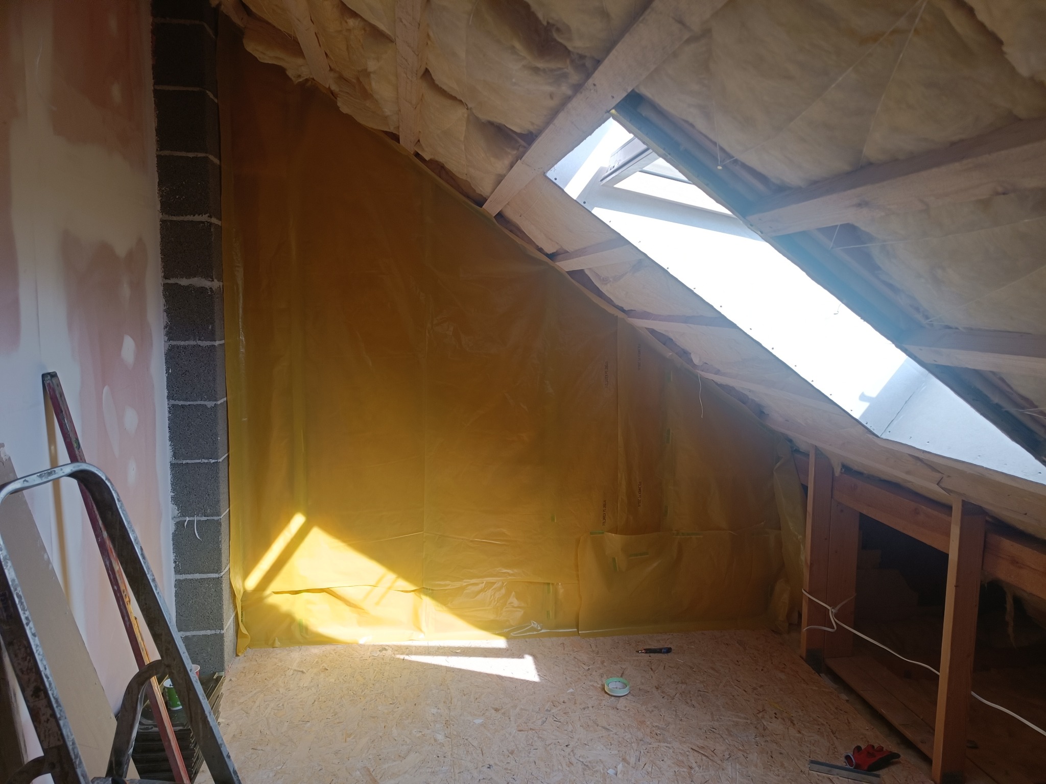 Poddasze w trakcie adaptacji: widoczna izolacja z wełny mineralnej, folia paroizolacyjna na ścianach, surowe drewniane elementy konstrukcyjne, okno dachowe wpuszczające silne światło, oraz drabina...