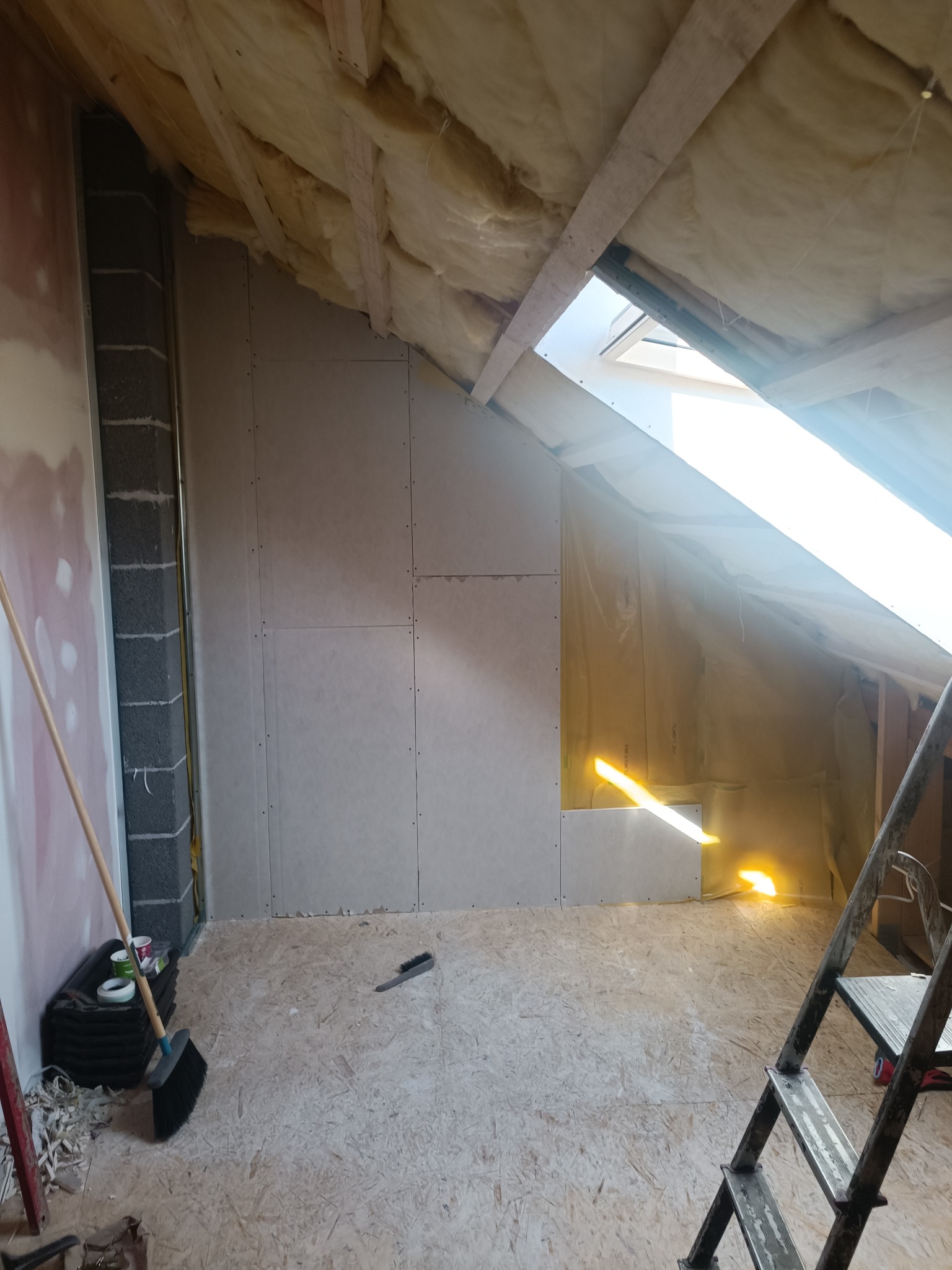 Poddasze w trakcie adaptacji: ocieplenie wełną mineralną, montaż płyt kartonowo-gipsowych na ścianach, widoczna drabina, narzędzia i resztki materiałów na podłodze OSB.
