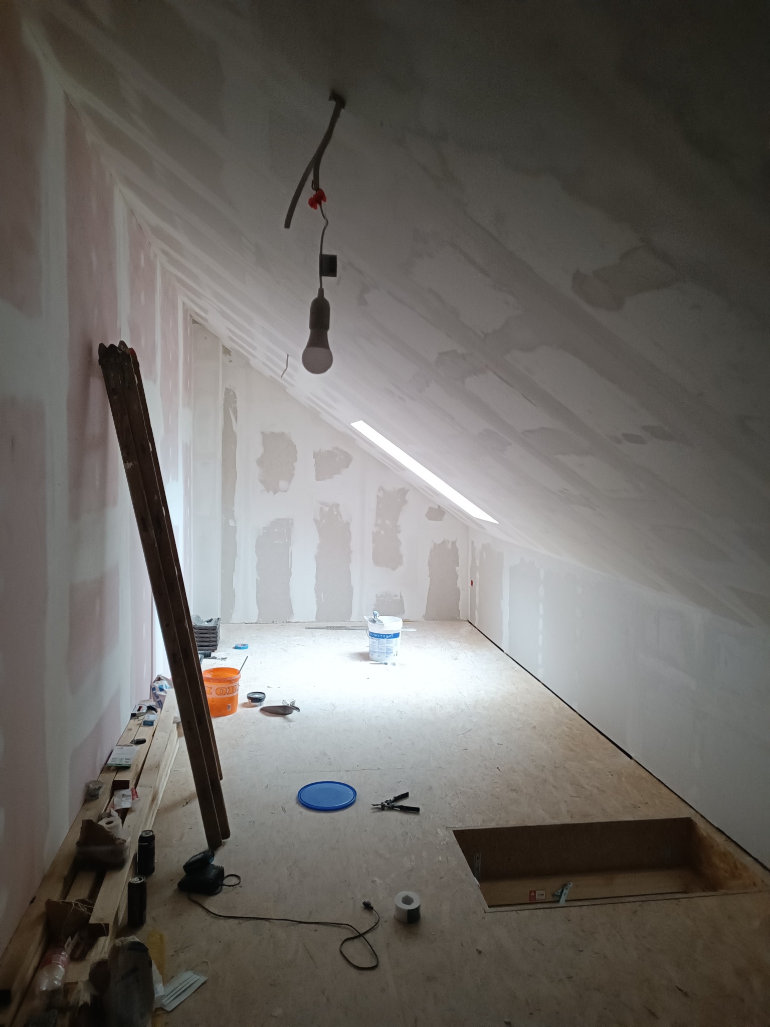 Poddasze w trakcie prac wykończeniowych: ściany i skosy pokryte płytami gipsowo-kartonowymi, częściowo wyszpachlowane, widoczne narzędzia i drabina oparta o ścianę, surowe oświetlenie żarówką.