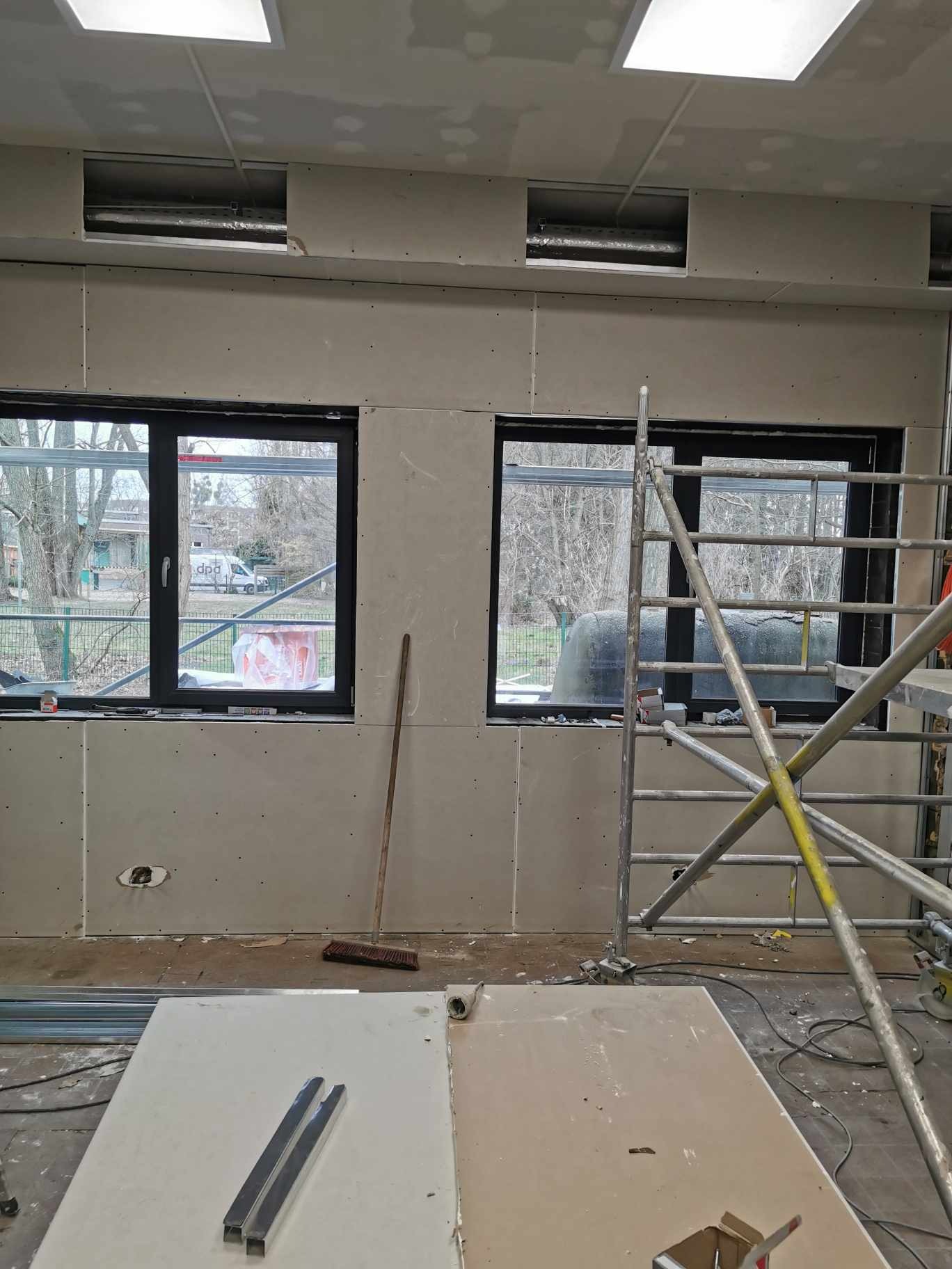 Wnętrze pomieszczenia w trakcie montażu płyt gipsowo-kartonowych na ścianach, widoczne okna z czarnymi ramami, rusztowanie, elementy konstrukcyjne na podłodze i otwory wentylacyjne w górnej części...