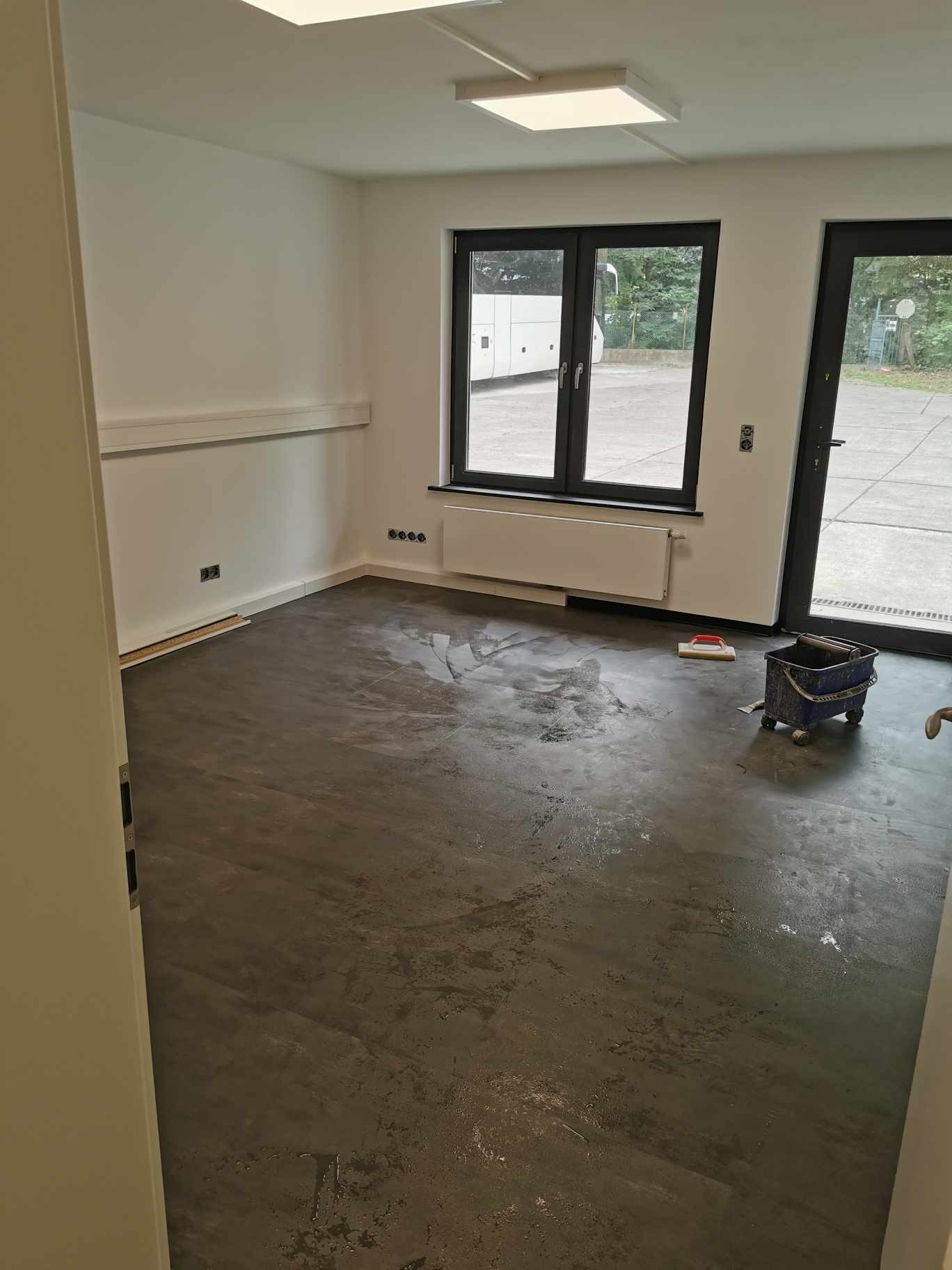 Przygotowanie podłoża pod posadzkę w biurze: mokra wylewka na ciemnej podłodze, wiadro z narzędziami w rogu, białe ściany i okno z widokiem na autobus.